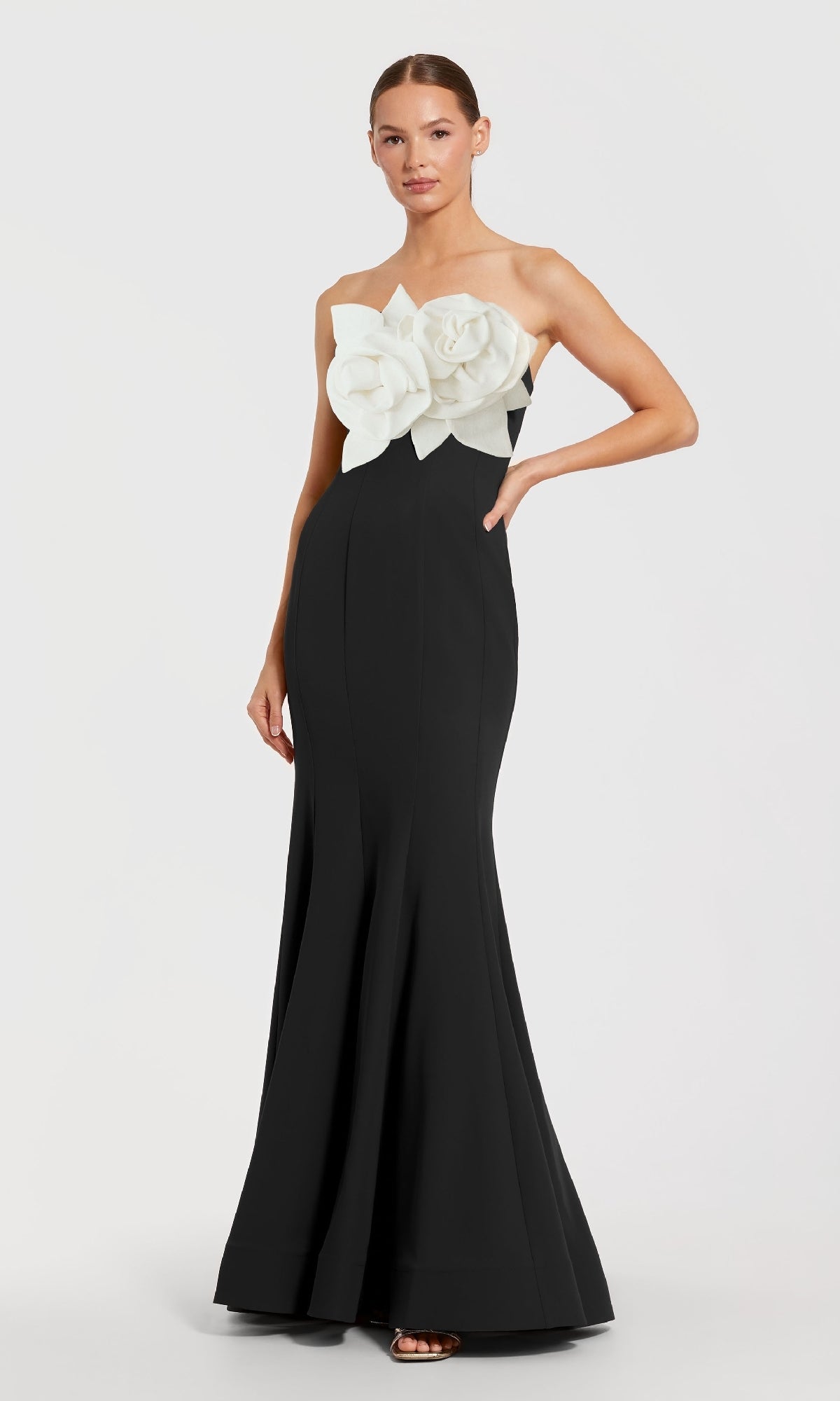 A woman stands against a plain background wearing the Mac Duggal 2300, a strapless long black formal dress featuring bold white floral appliqués on the bodice.